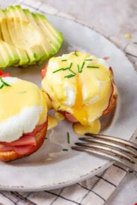 A white plate with the whole eggs Benedict and half of an eggs Benedict. There is a sliced avocado behind the eggs Benedict.