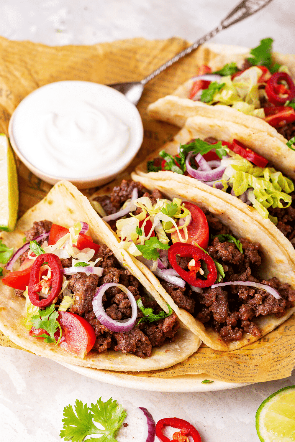 Two whole ground beef tacos and part of two ground beef tacos on a piece of newspaper on a white plate. Behind the tacos is a small cup of sour cream.