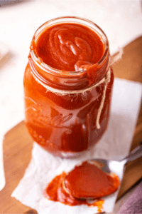 A glass jar filled all the way to the top with barbecue sauce. The jar is on a paper towel there's a spoon in front of the jar covered in barbecue sauce.