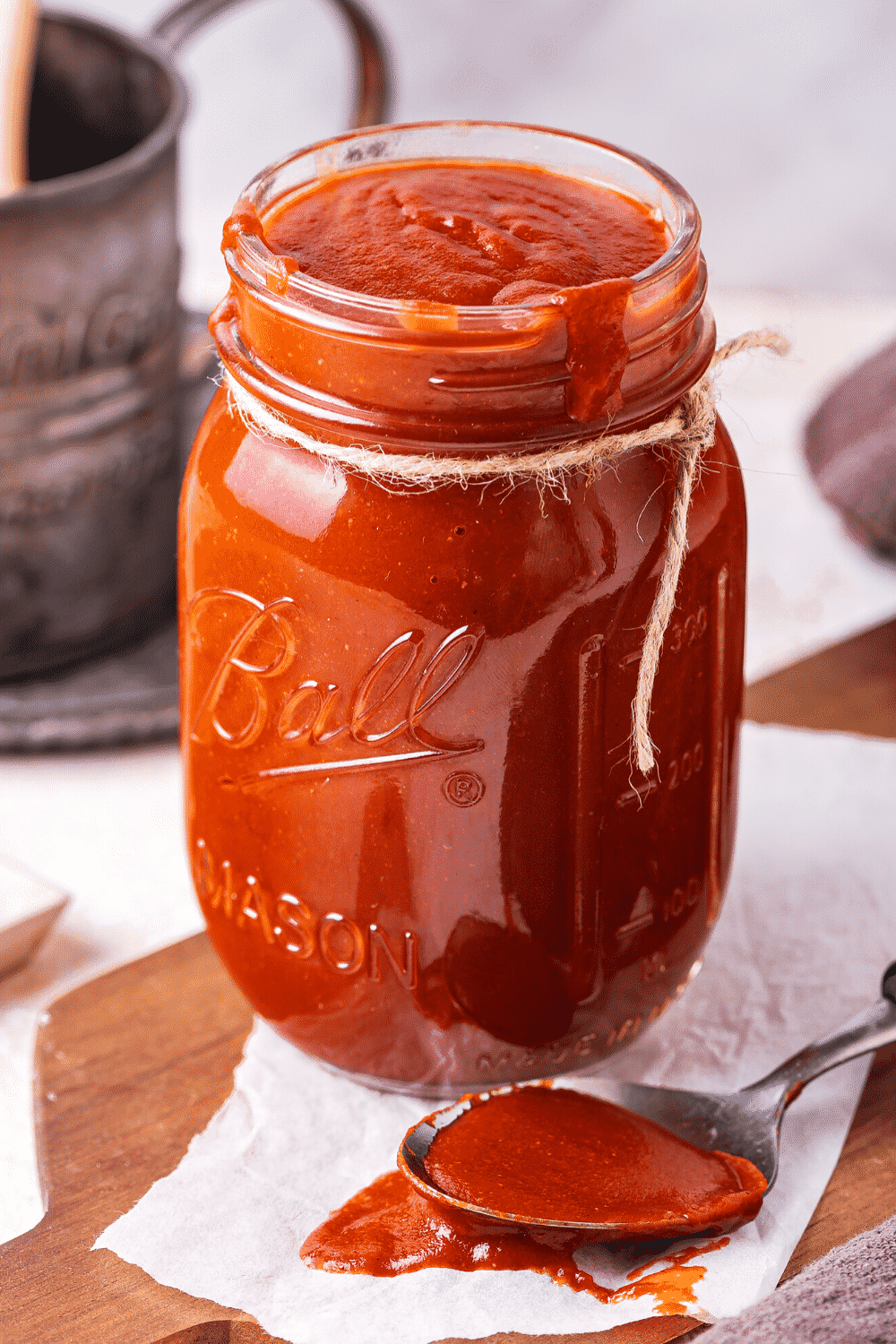 A glass jar filled to the top with barbecue sauce. The jars on a paper towel and there is a spoons filled with barbecue sauce in front of the jar on the paper towel.
