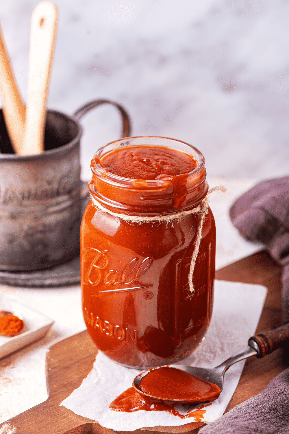 A glass jar filled with barbecue sauce on a paper towel on a wooden cutting board. There is a spoon with barbecue sauce on it on the paper towel.