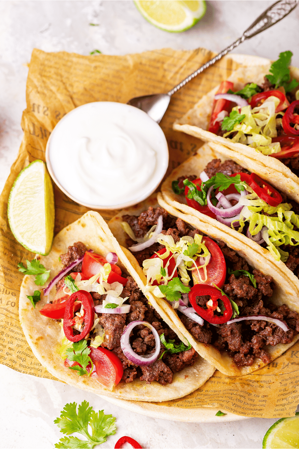 Three ground beef tacos next to one another with part of a ground beef taco at the back end of them. A cup of sour cream is behind the three tacos.