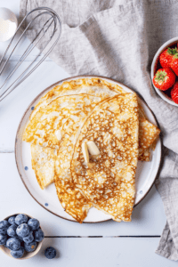 A white plate with four crepes overlapping one another on it. There is a small bowl of blueberries in front of the plate and a small bowl of strawberries behind the plate.