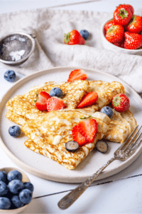 A white plate with four crepes overlapping one another on it. There are slices of strawberries and blueberries on the crepes and there is a bowl of blueberries in front of the plate and a small bowl of strawberries behind the plate.