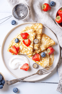 A white plate with four crepes on it. There are slices of blueberries and strawberries on the crepes.