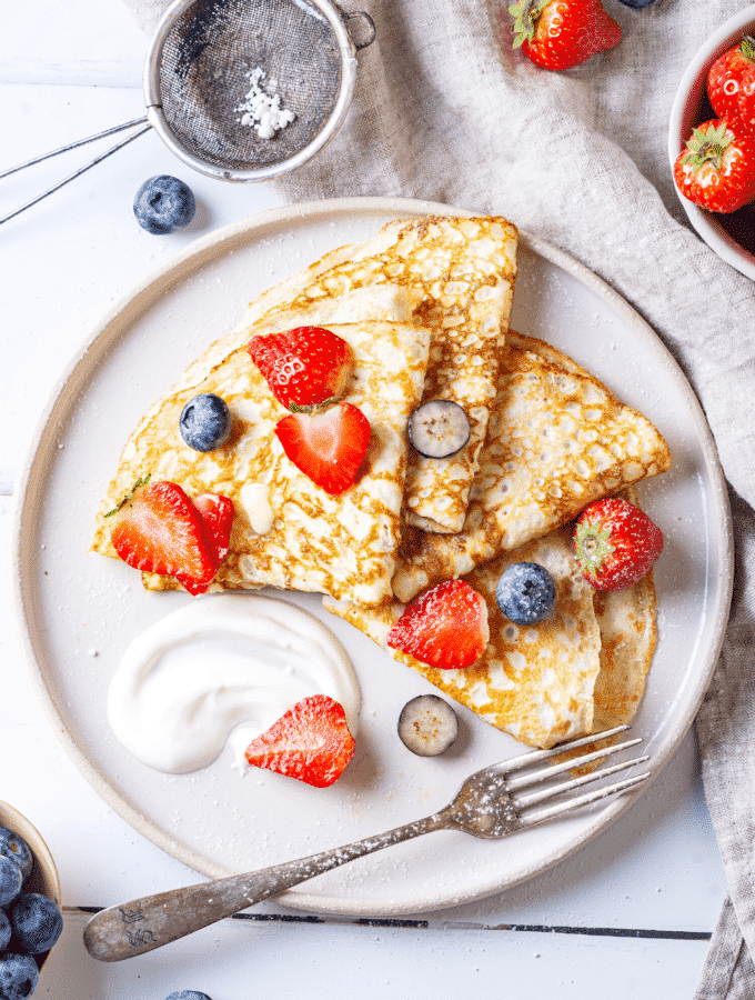 A white plate with four crepes on it. There are slices of blueberries and strawberries on the crepes.