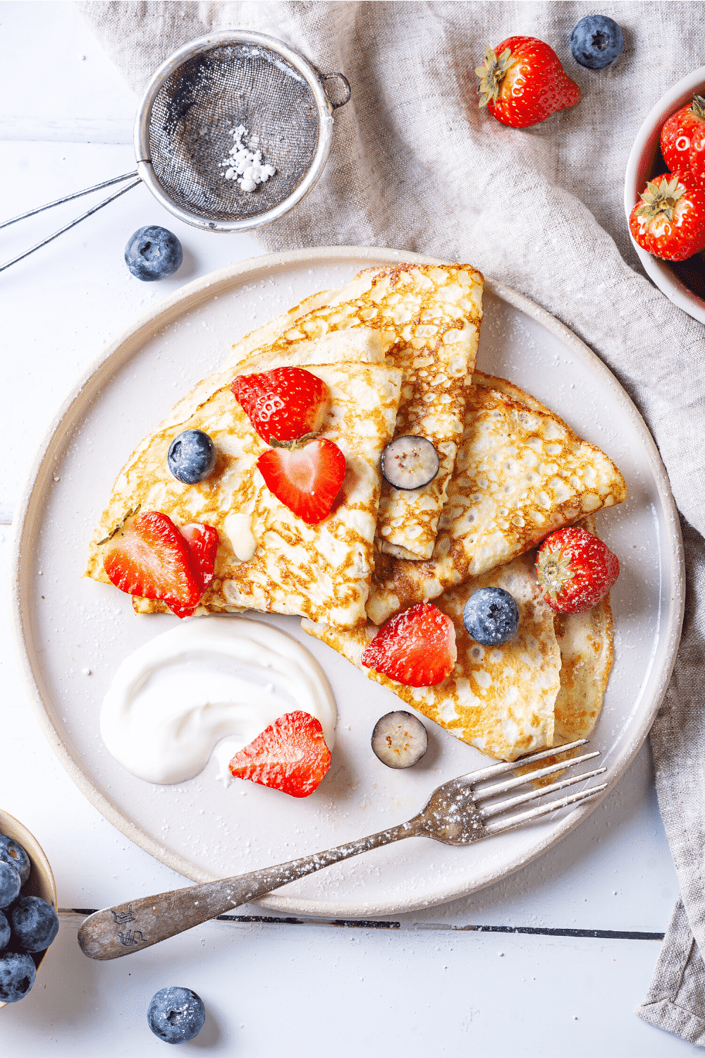 A white plate with four crepes on it. There are slices of blueberries and strawberries on the crepes.