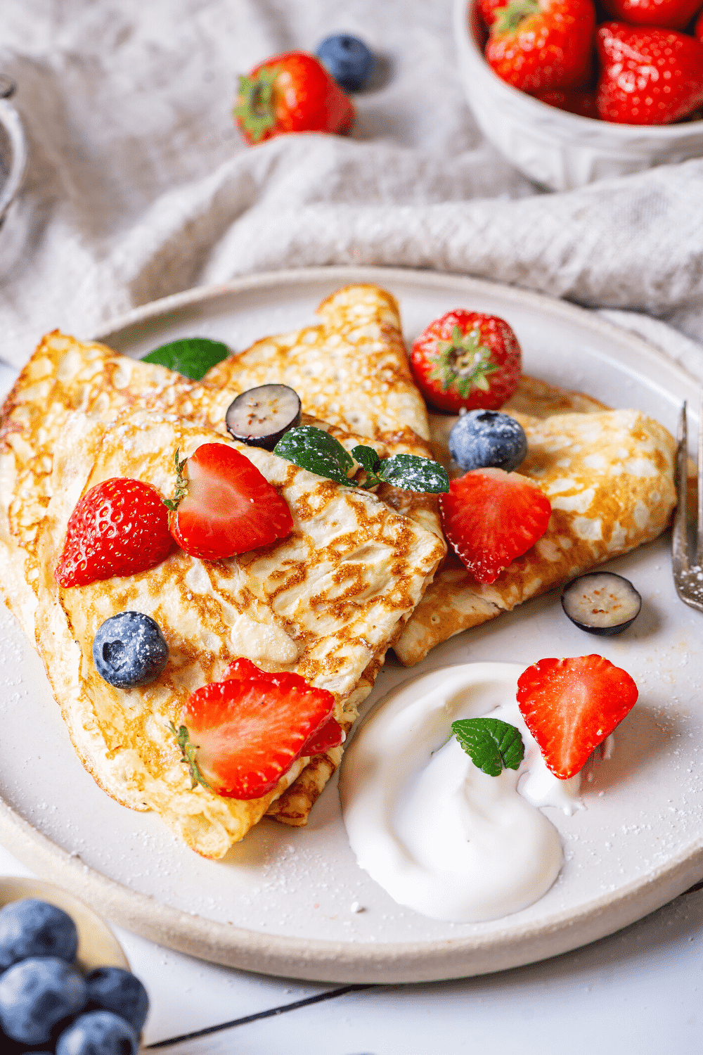 A white plate with a few crepes on it. There are slices of strawberries and a few blueberries on the crepes.