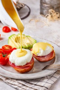 A white plate with two eggs Benedict on it. A gravy boat is pouring hollandaise sauce on one of the eggs. There are sliced grape tomatoes behind that egg and a sliced avocado behind the other egg.