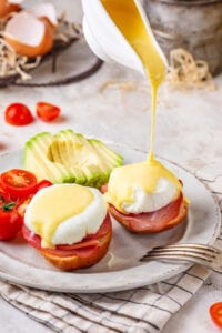 A white plate with two eggs Benedict on it. A gravy boat is pouring hollandaise sauce over top of the one eggs Benedict. There is a sliced avocado behind it and grape tomatoes to the left of the avocado.