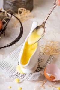 A gravy boat filled with hollandaise sauce on a piece of newspaper. A spoon is hovering over the boat with some hollandaise on it dripping down into the boat.