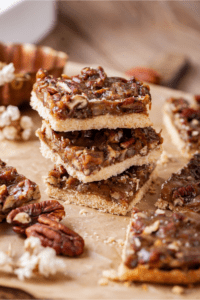 Three pecan pie bars stacked on top of one another on a piece of parchment paper. There are parts of three pecan pie bars to the right of the stack.