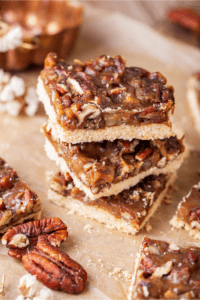 Three pecan pie bars stacked on top of each other on a piece of parchment paper. There are parts of other pecan pie bars and pecans around the stack.