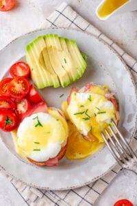 A white plate with two eggs Benedict, a couple of sliced grape tomatoes, and a sliced avocado on it.