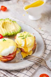 A white plate with two eggs Benedict on it. A sliced avocado is on the plate behind the eggs benedict.