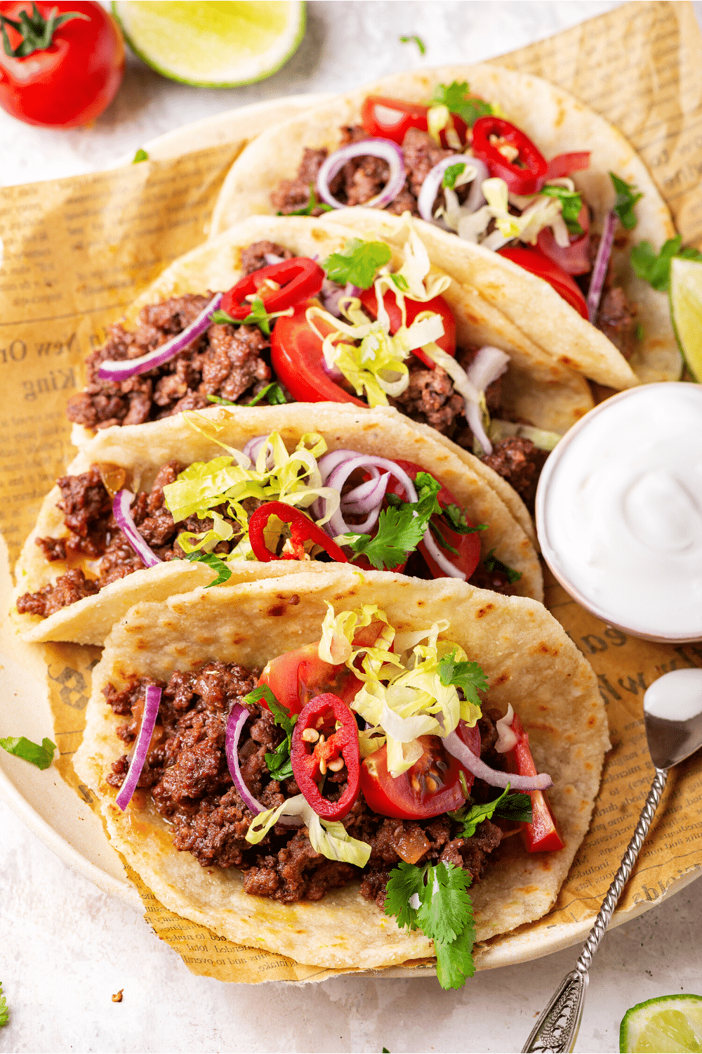 For ground beef tacos in a row on a piece of newspaper. A small cup of sour cream is to the right of the middle tacos.