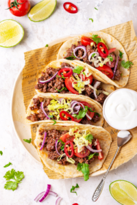 Four ground beef tacos in a row on a piece of newspaper on a plate. There is a small cup of sour cream to the right of the tacos.