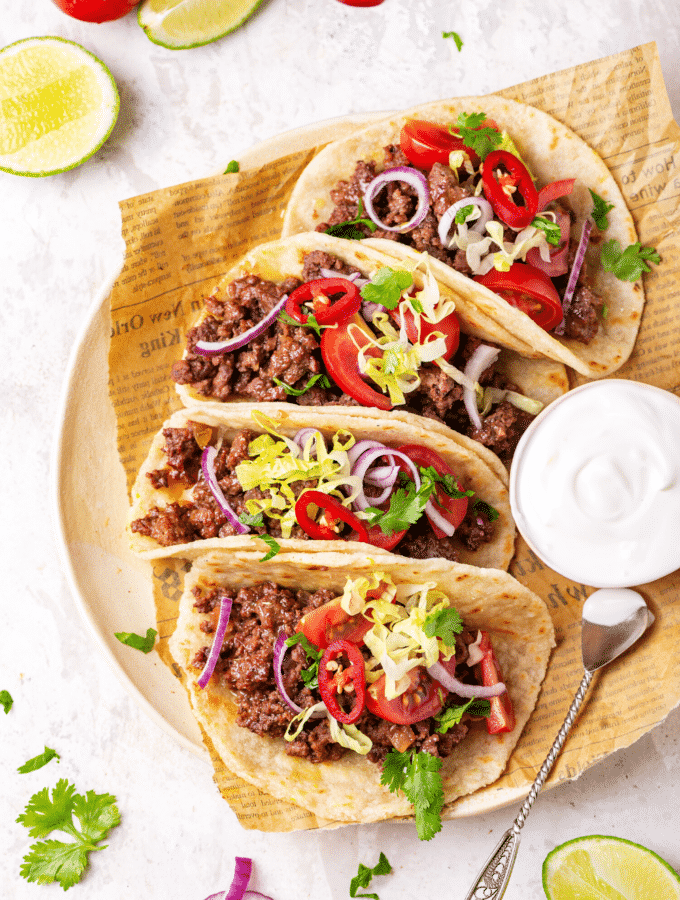 Four ground beef tacos in a row on a piece of newspaper on a plate. There is a small cup of sour cream to the right of the tacos.