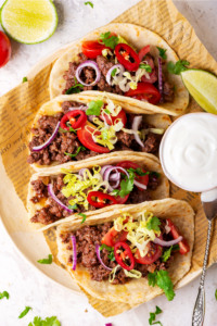 A row of four ground beef tacos on a piece of newspaper. To the right of the two middle tacos it's a small cup of sour cream.