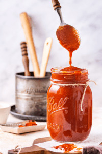 A glass jar filled to the top with barbecue sauce. A spoon is hovering over the jar dripping barbecue sauce down from it into the top of the jar.