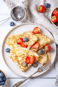 For crêpes overlapping one another on a white plate. They are slices of strawberries and blueberries on top of the crepes.