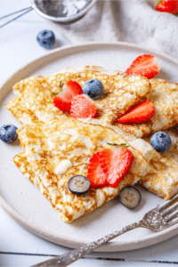 A white plate with a few crepes on it with some strawberry and blueberry slices on top.