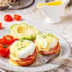 Two eggs benedict on a white plate with sliced tomatoes and a sliced avocado behind it. There is a gravy boat filled with hollandaise sauce behind it.