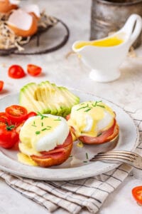 Two eggs benedict on a white plate with sliced tomatoes and a sliced avocado behind it. There is a gravy boat filled with hollandaise sauce behind it.