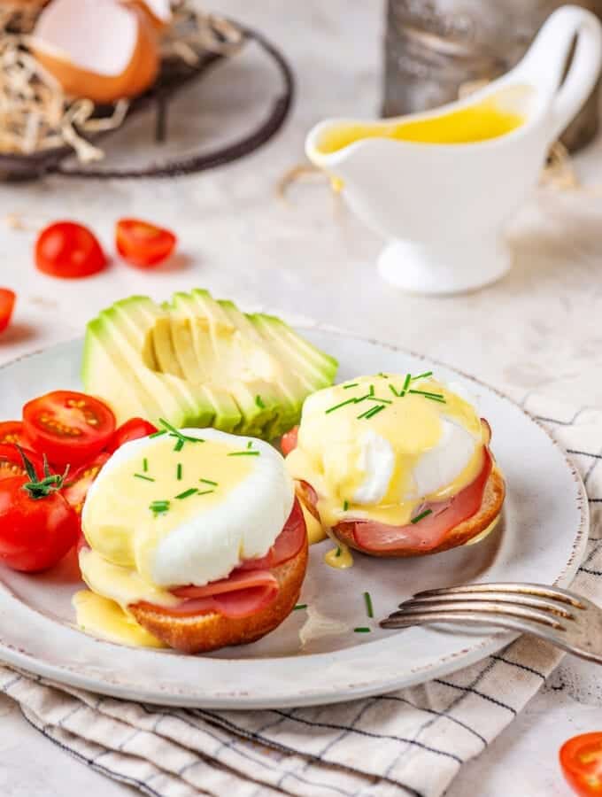 Two eggs benedict on a white plate with sliced tomatoes and a sliced avocado behind it. There is a gravy boat filled with hollandaise sauce behind it.