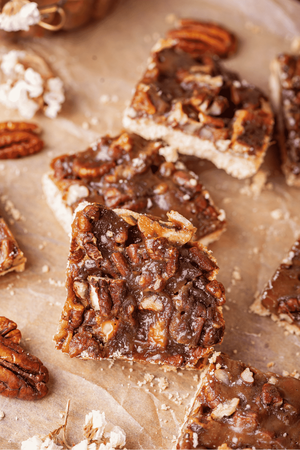 Three pecan pie bars overlapping one another on a piece of parchment paper. There are parts of pecan pie bars to the right and pecans to the left.