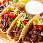 Three ground beef tacos in a horizontal row on a piece of newspaper on a white plate. Small cup of sour cream is behind tacos.