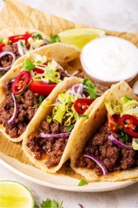 Three ground beef tacos in a horizontal row on a piece of newspaper on a white plate. Small cup of sour cream is behind tacos.