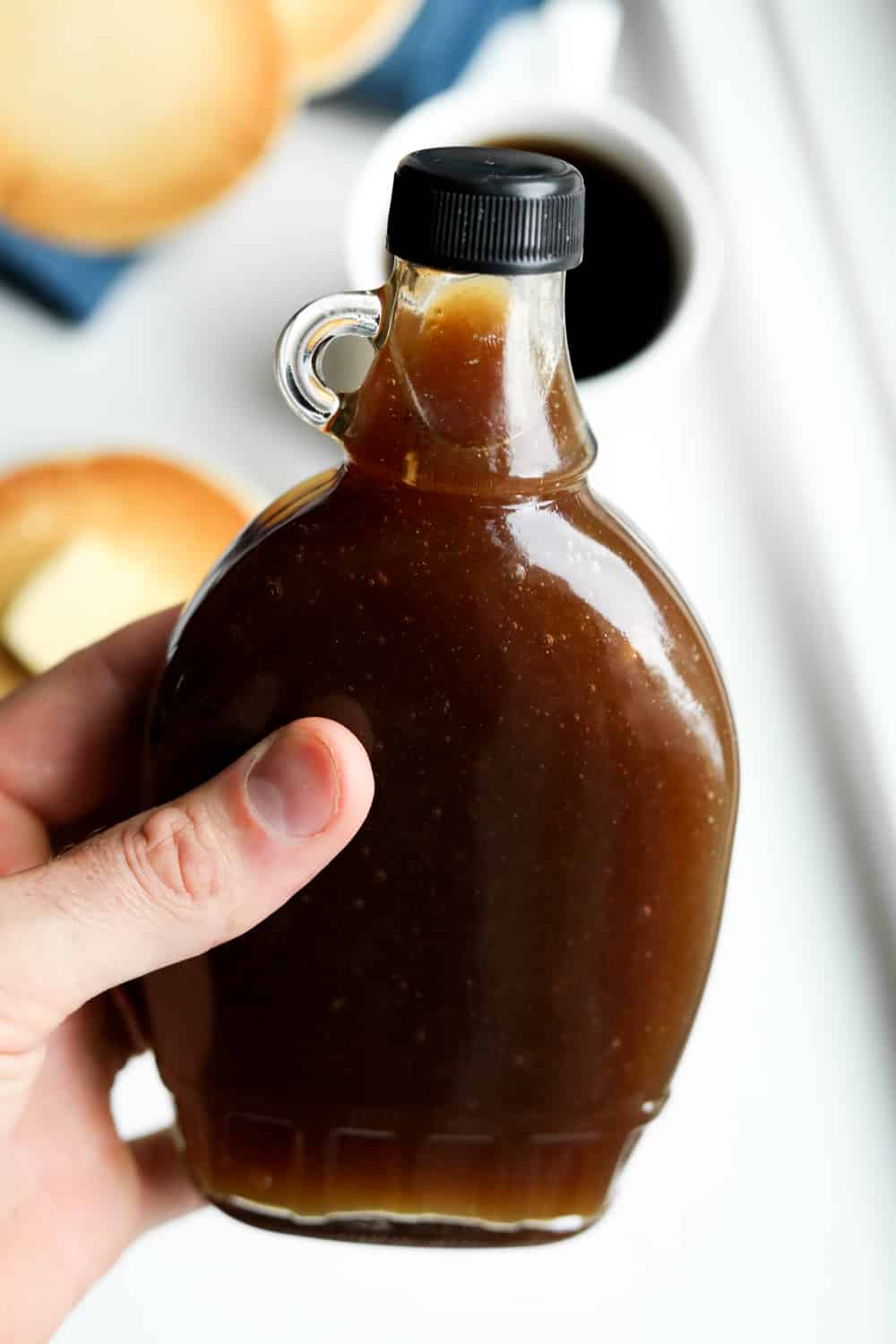 A hand holding a bottle of maple syrup over a plate of pancakes.