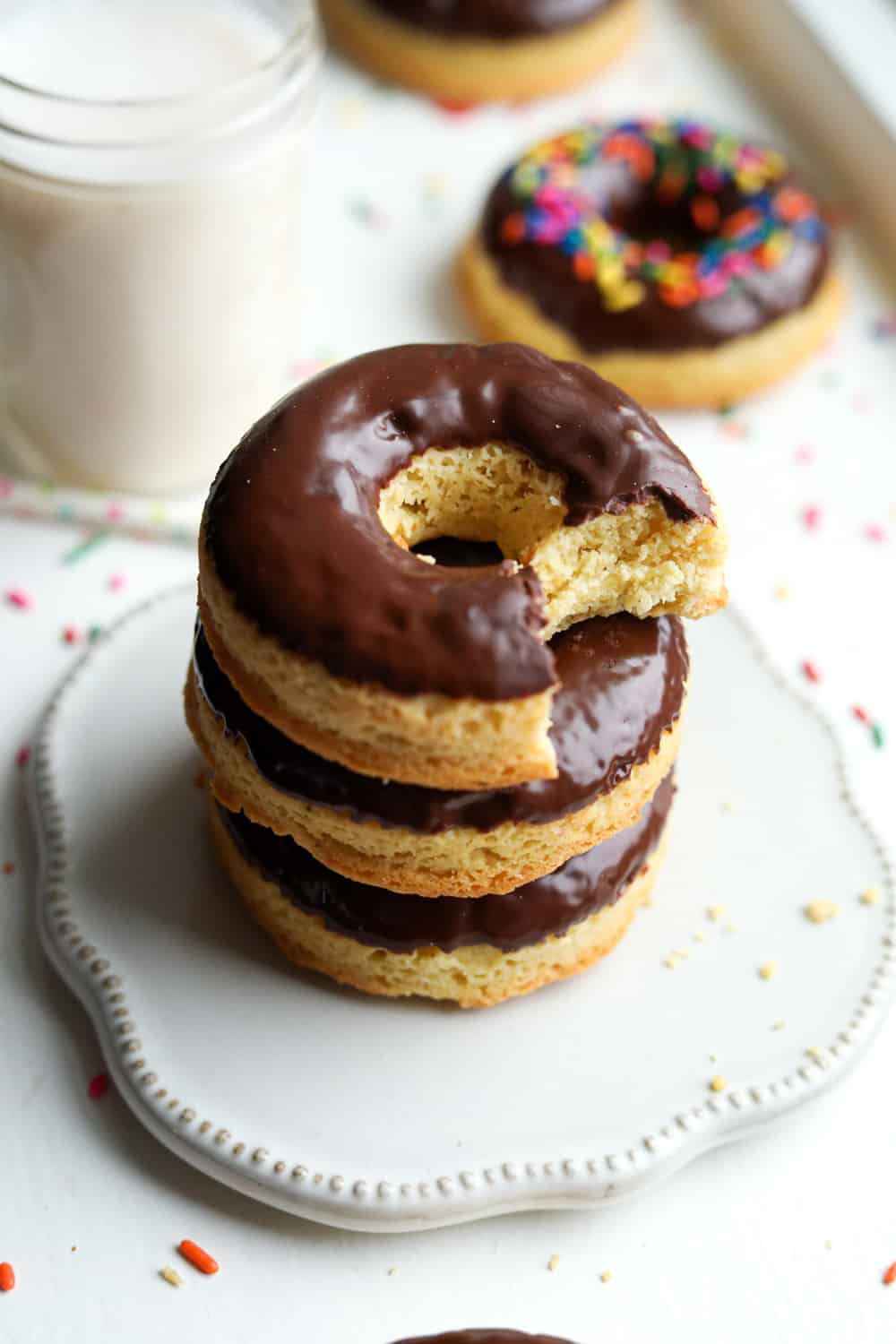 Three donuts stacked on top of each other on a serving dish. There's a glass of milk and other donuts behind them.