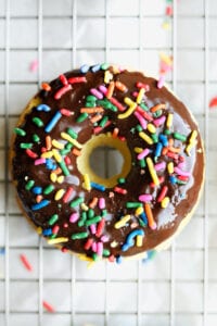 A chocolate covered donut topped with multi-colored sprinkles on a silver wire rack.