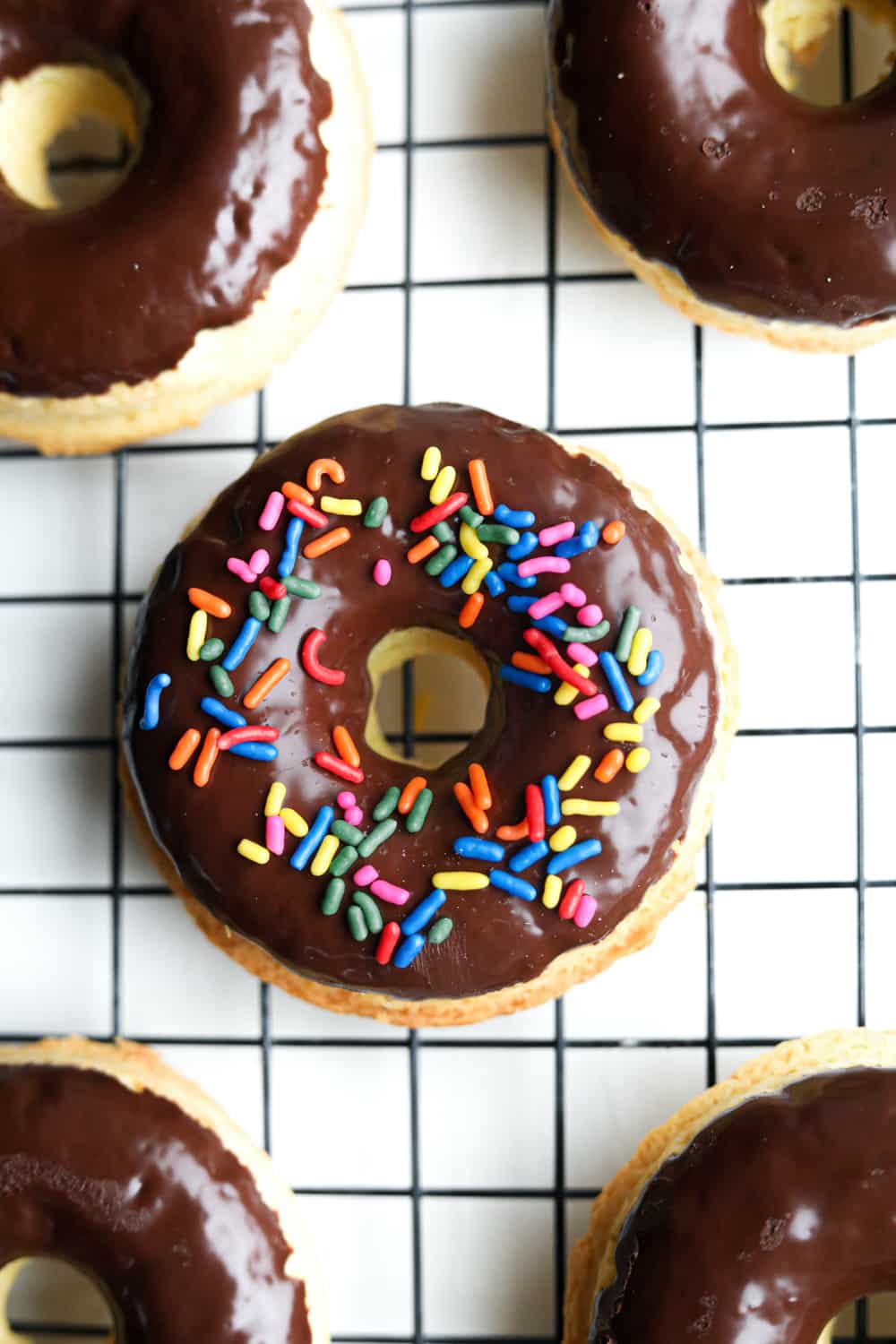 A chocolate covered donut that has been topped with sprinkles on the drying rack. There are other donuts set next to it.