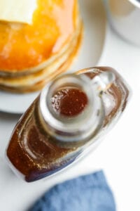 A bottle of maple syrup next to a plate of pancakes.