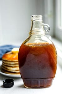 A bottle of maple syrup in front of a plate of pancakes.