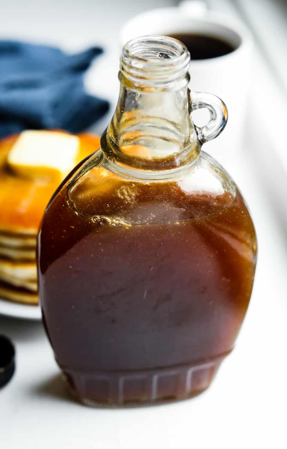 A bottle of maple syrup sat in front of a plate of pancakes, a blue napkin, and a cup of coffee.