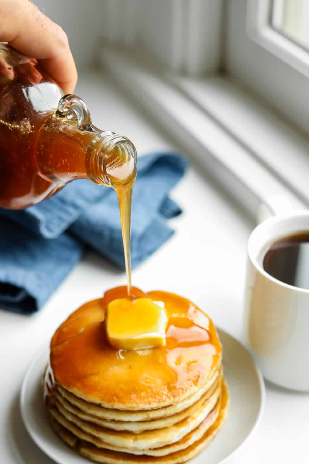 A hand holding a bottle of maple syrup and pouring it over a stack of pancakes.