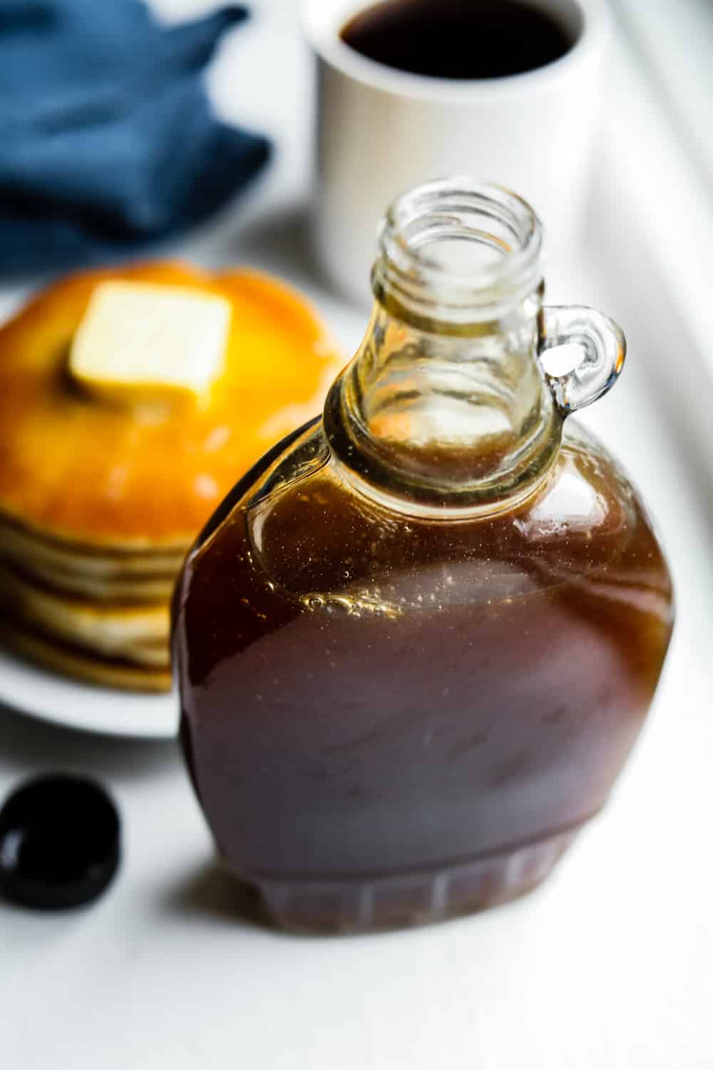 A bottle of maple syrup in front of a plate of pancakes and a blue napkin.