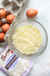 A bowl of cheese, brown eggs, and xanthan gum on a white table.