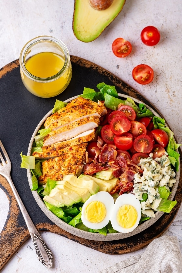 A plate of carbs salad on a serving board. There is a fork to the left of the plate and a glass jar filled with Dijon vinaigrette dressing. A whole cherry tomato a cherry tomato cut in half and part of an avocado are behind the serving tray.