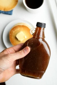 A hand holding a bottle of maple syrup over a plate of pancakes and a cup of coffee.