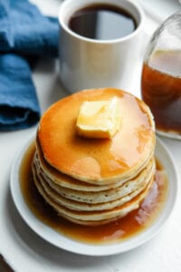 A stack of pancakes covered in maple syrup. There's a blue napkin a cup of coffee and a bottle of syrup behind the pancakes.