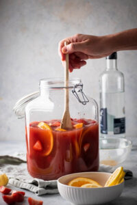 A glass gallon jar filled ¾ of the way with jungle juice. Hand is holding a wooden spoon mixing the jungle juice. In front of it is a small white bowl orange slices and behind it a bottle of vodka