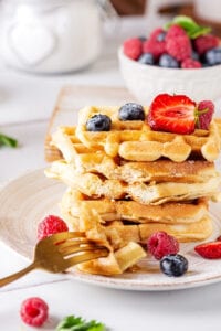 Stack of five waffles on a white plate. There is a fork sat on the edge of the plate with part of two waffles on the prongs. A few raspberries and blueberries are on the waffle and plate and there is a bowl of raspberries and blueberries behind the plate.