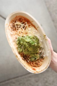 A hand holding a keto chipotle bowl with Barbacoa, sour cream, shredded cheese, and guacamole..