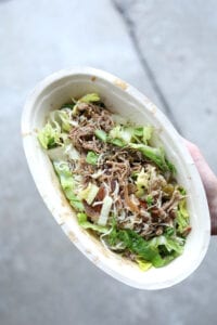 A hand holding a keto chipotle bowl with Carnitas, Romain lettuce, and shredded cheese.
