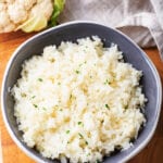 A gray bowl filled with cauliflower rice on top of the wooden cutting board. Part of a cauliflower head and part of a gray napkin are behind the bowl.